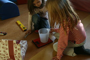 zwei Mädchen wiegen einen Haushalts-Messbecher auf einer dünnen roten Elektrowaage